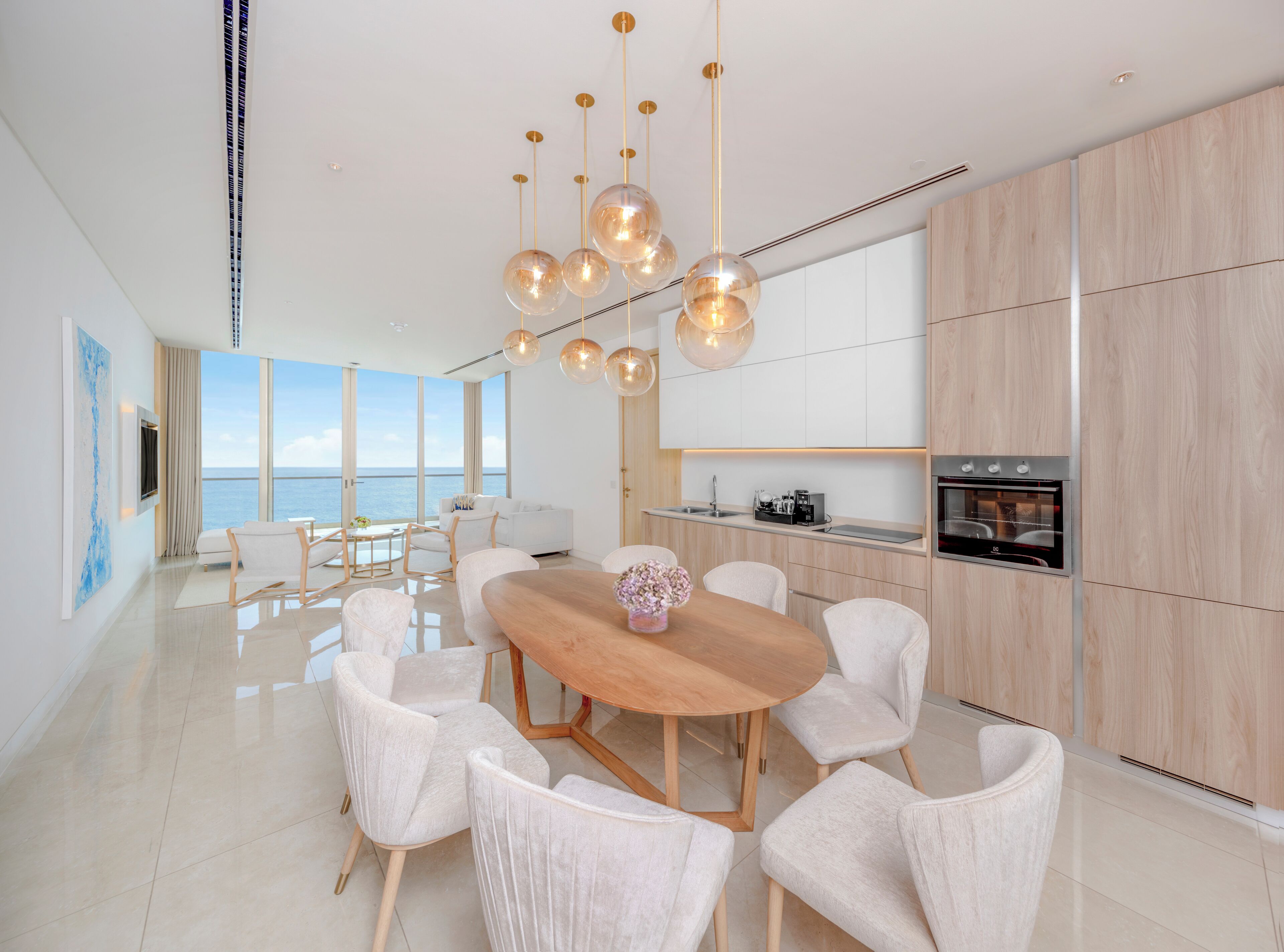 A modern kitchen with a central island, a dining area with a wooden table and white chairs, and a large window offering a view of the sea.