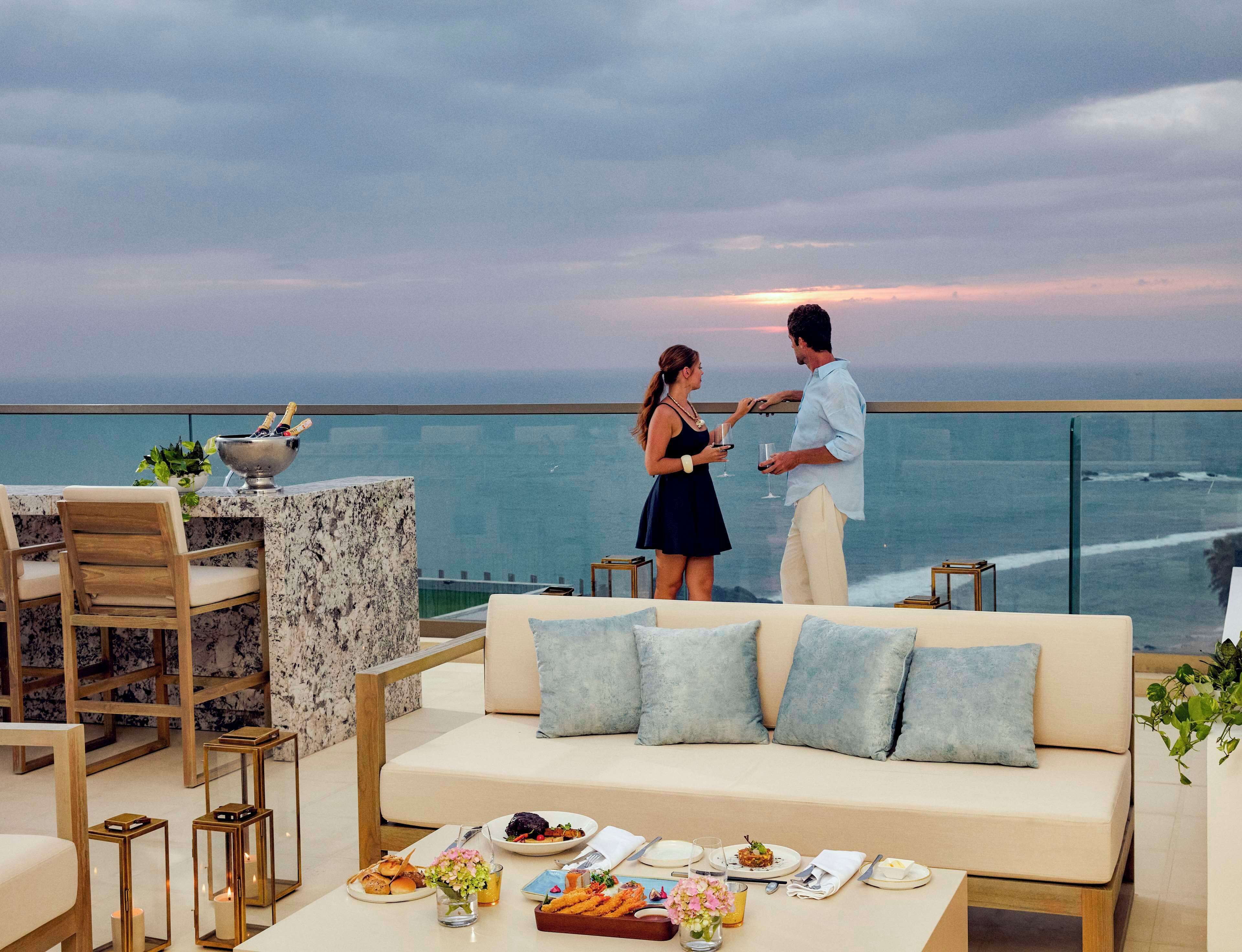 A couple dining on a balcony with a view of the ocean, a marble bar, and a sofa with cushions.