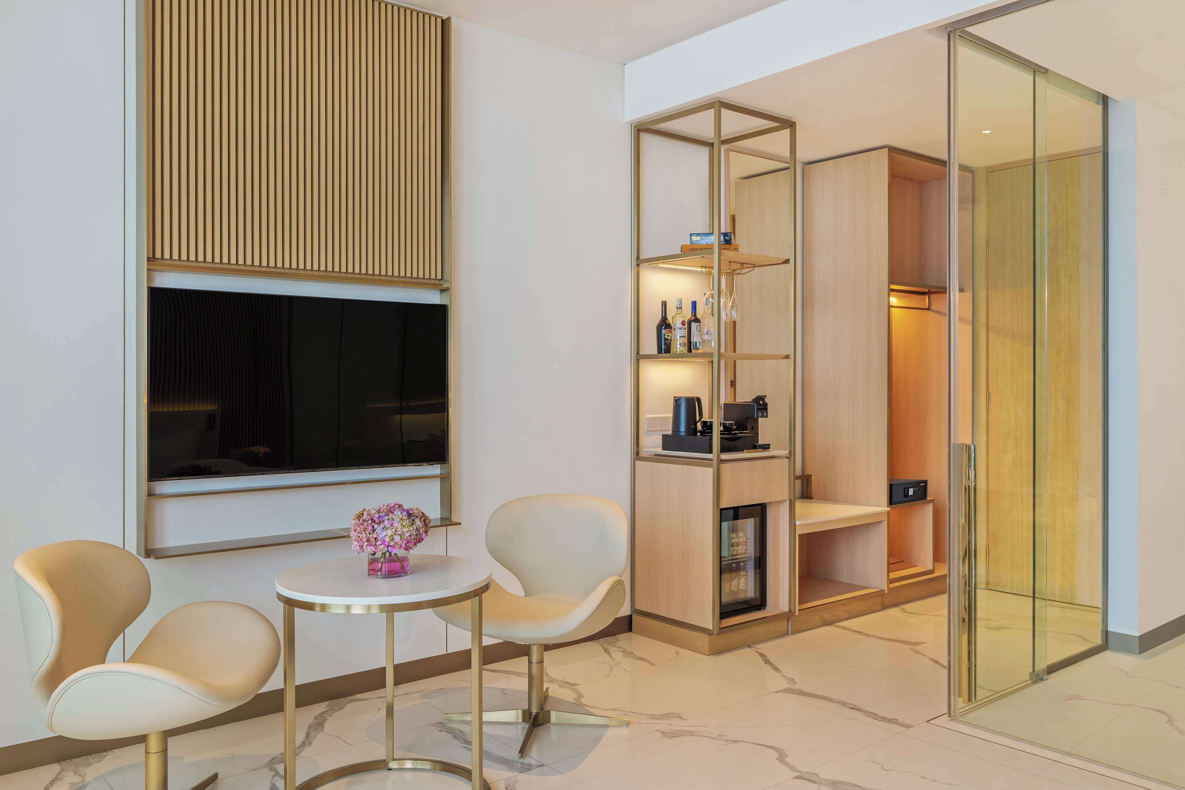 A modern hotel room with a seating area, a large window with blinds, a built-in kitchen, and a glass-enclosed closet.