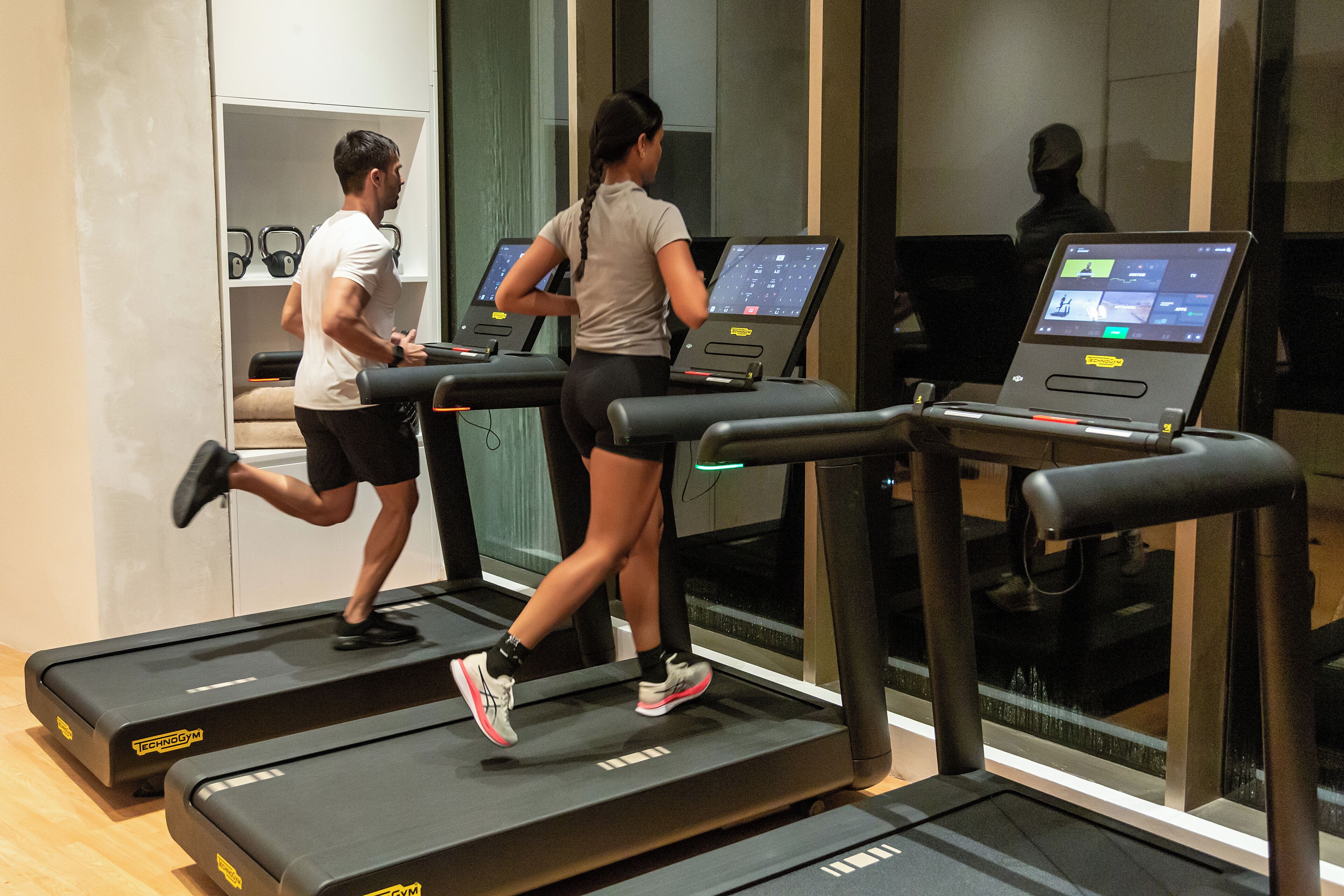 Two people running on treadmills in a gym with a mirrored wall and a shelf with kettlebells.