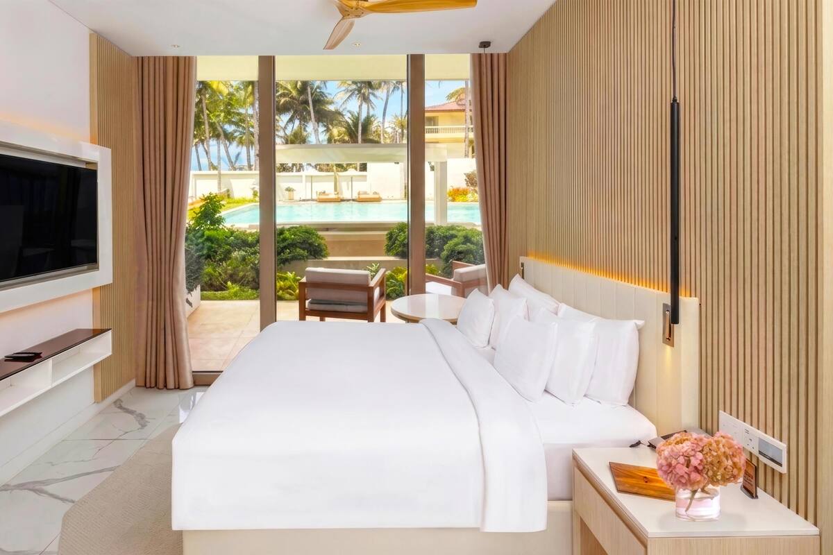 Une chambre d’hôtel moderne avec un grand lit, un téléviseur à écran plat, une petite table avec un vase, et une vue sur une piscine et des palmiers.
