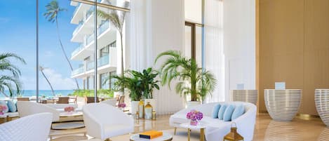A spacious, modern lounge area with large windows, white furniture, and potted plants.