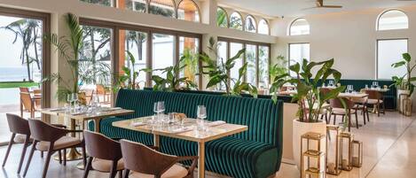 A restaurant with a green cushioned seating area, multiple tables with chairs, and large windows with a view of the ocean.