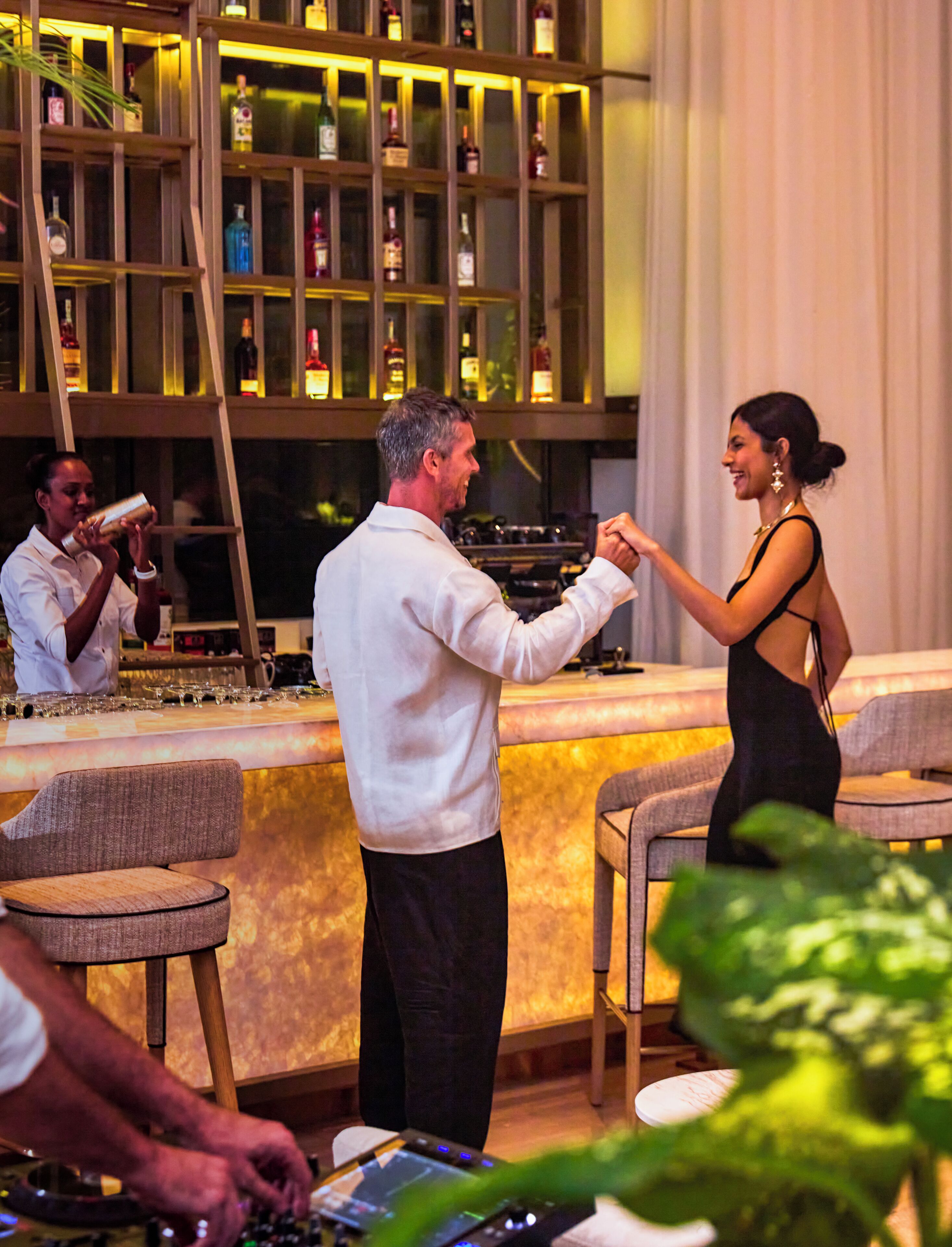 A bar area with a bartender, a woman in a black dress, and a man in a white shirt. There are shelves with bottles in the background.
