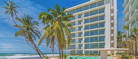 A modern hotel with a pool area, palm trees, and a beach view.