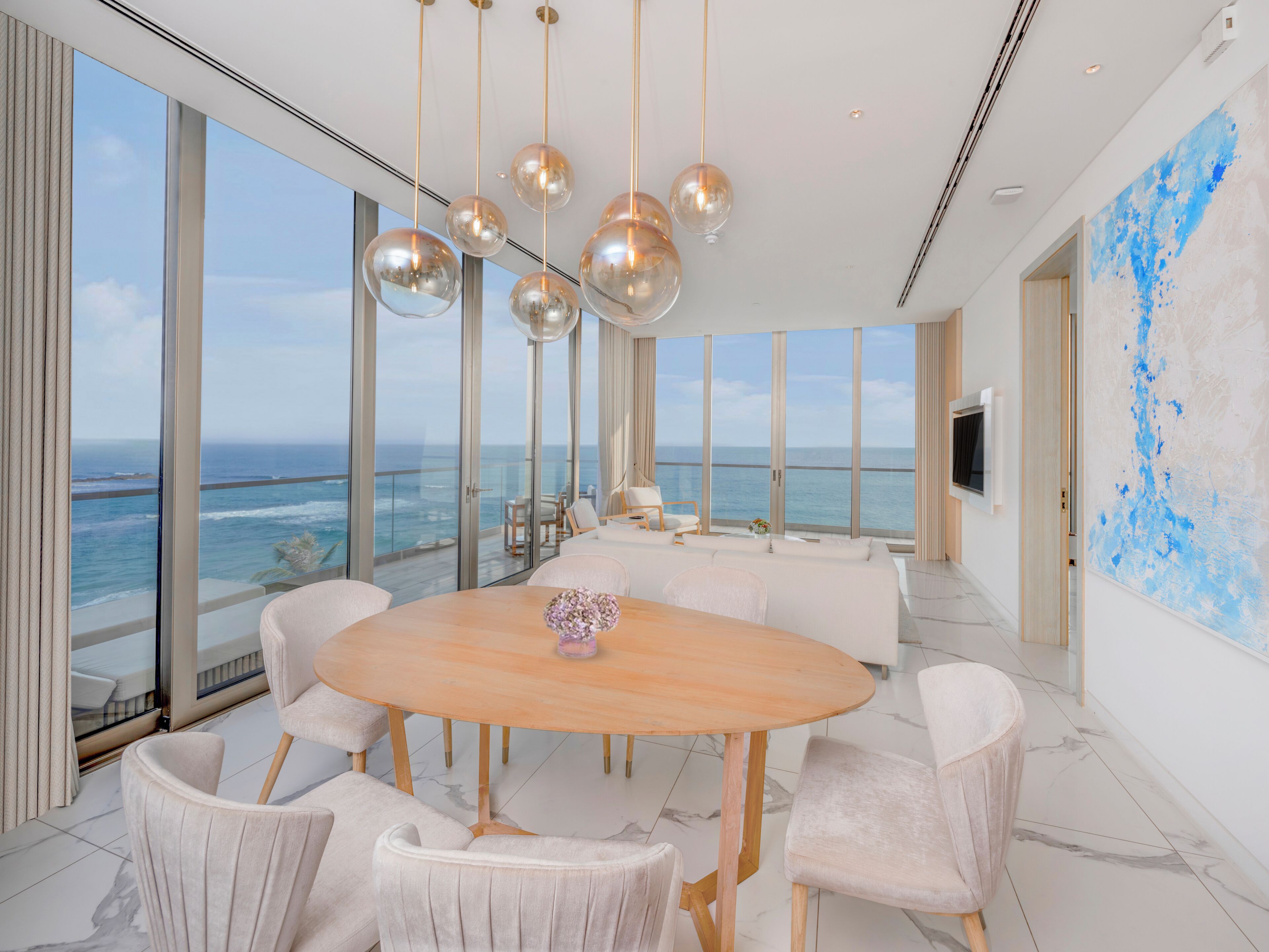 Un espace repas moderne avec une grande table ronde, des chaises à revêtement blanc et une vue sur l’océan grâce à de grandes fenêtres du sol au plafond.