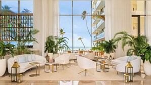Amplia sala de estar con un ventanal que da a una playa, amoblada con sofás y sillones blancos, rodeada de plantas en macetas.