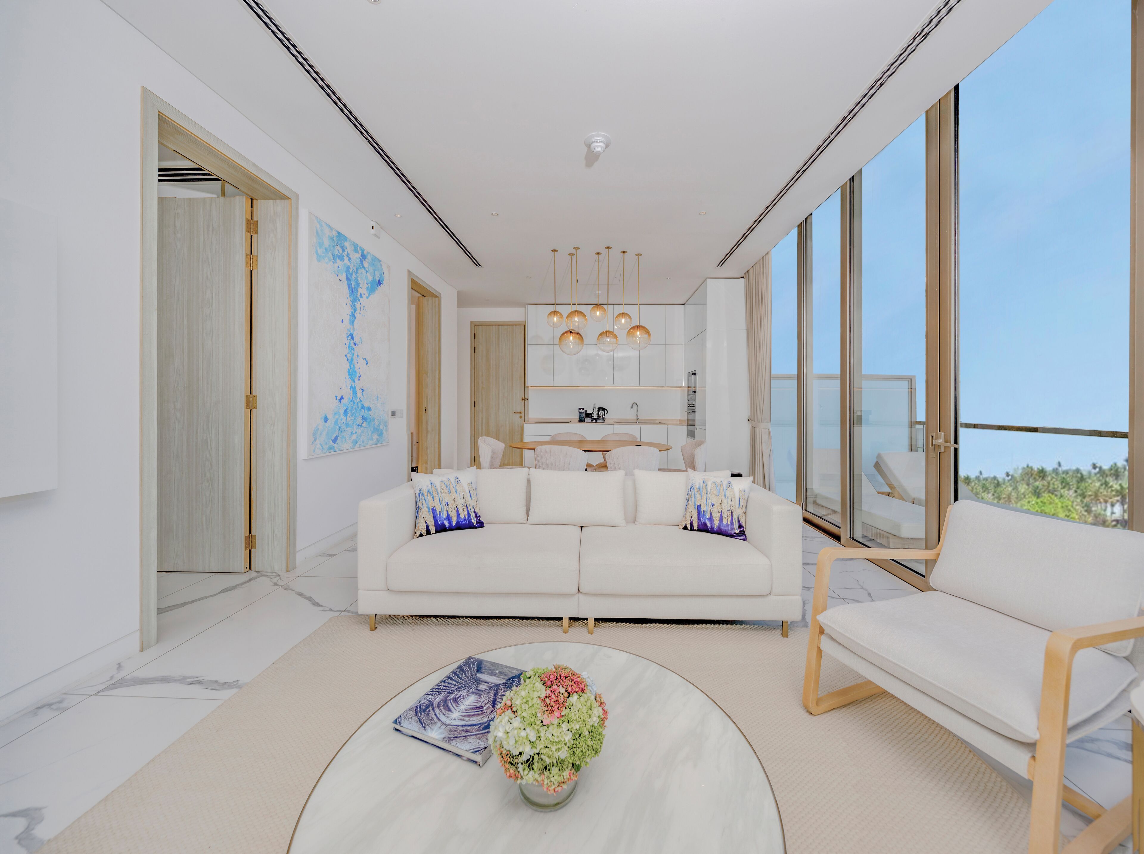A modern living room with a white sofa, a coffee table with a book and flowers, and a balcony with a view.