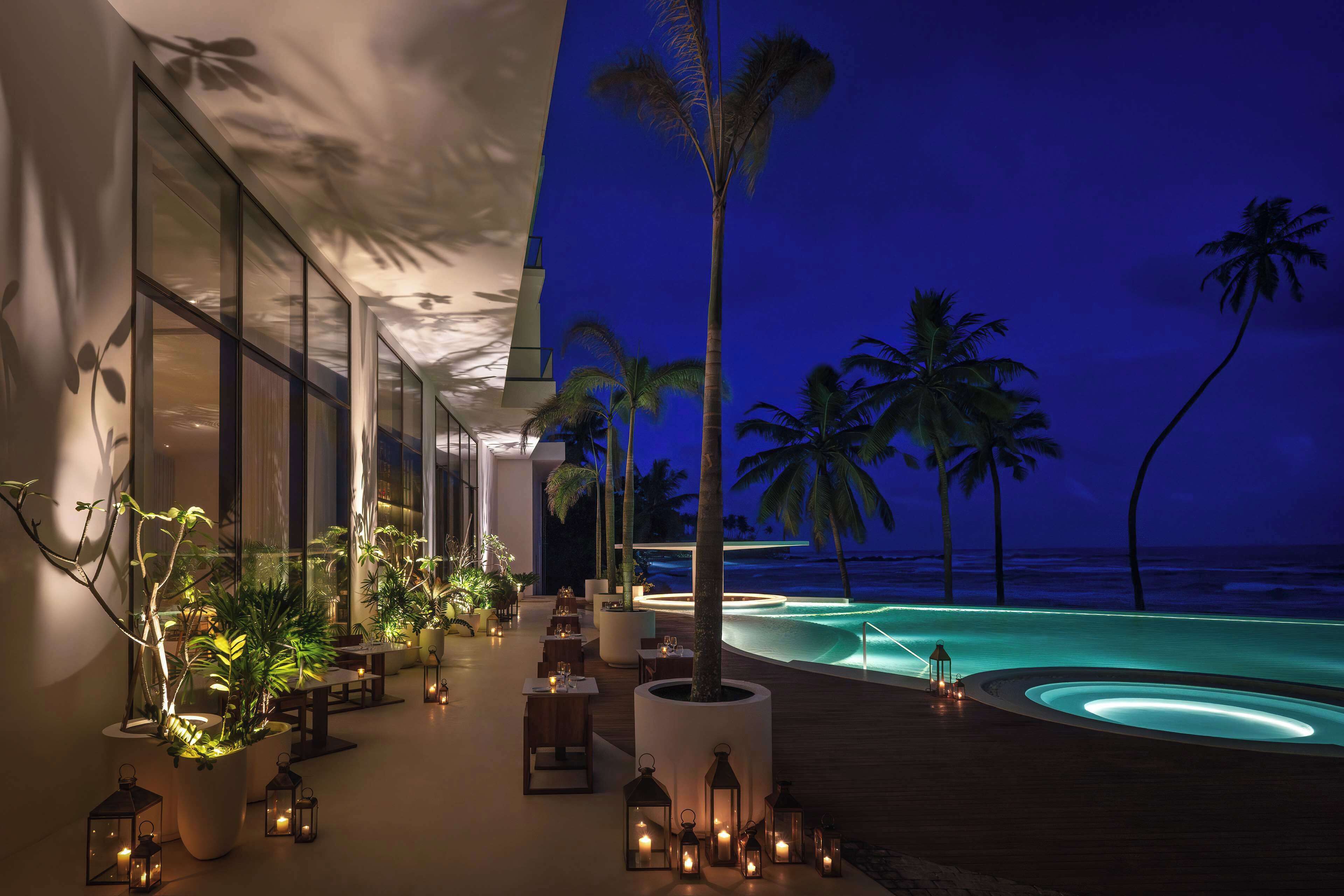 A resort pool area with a view of the ocean at night, featuring palm trees, outdoor seating, and ambient lighting.