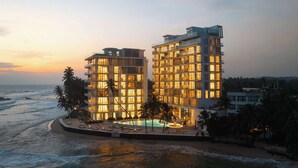 Un hotel moderno de gran altura con varios balcones y zona de piscina, cerca de una playa con vistas al océano.