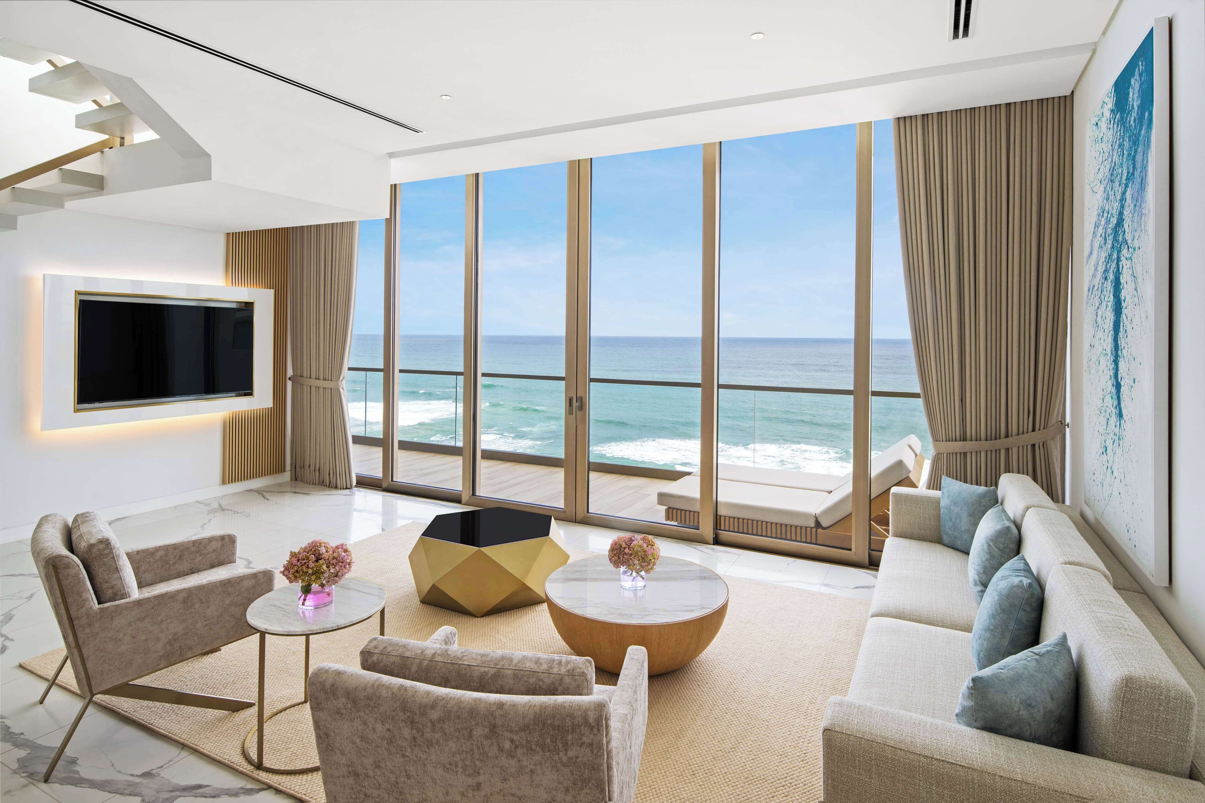 A modern living room with a large window overlooking the ocean, a flat-screen TV mounted on the wall, and a comfortable seating area with sofas and a coffee table.