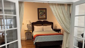Une chambre  à coucher avec un lit en bois, deux tables de chevet, une commode, un tableau encadré au mur et une fenêtre avec des rideaux.