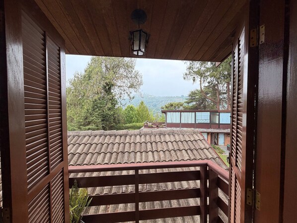 A wooden balcony with a tiled roof, overlooking a mountainous landscape and a building with a blue facade. - THE WRITERS HOUSE (Campos do Jordão)