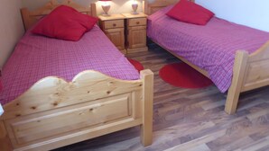 Een kamer met twee houten bedden, elk met een rode kussen en een geruit plaid. Twee nachtkastjes met lampen en een klein houten kastje tussen de bedden.