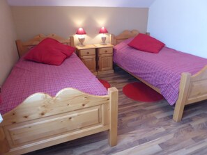 Een kamer met twee houten bedden, elk met een rode kussen en een geruit plaid. Twee nachtkastjes met lampen en een klein houten kastje tussen de bedden.