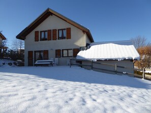 A two-story house with a sloped roof covered in snow, a wooden fence, and a snow-covered lawn.