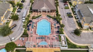 Vista aérea de una zona residencial con una gran piscina rodeada de sillones de descanso y sombrillas, un muelle que se extiende sobre el agua, y un edificio central con techo a dos aguas.