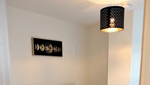 Un lit bien fait, avec du linge de lit blanc, une tête de lit sombre et une petite table de chevet surmontée d’une lampe. Au-dessus du lit, on peut voir une œuvre d’art encadrée représentant les différentes phases de la lune.