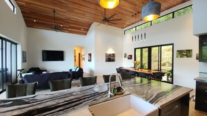 A modern living room with a large TV, comfortable seating, a kitchen area with a marble countertop, and large windows offering a view of greenery. - Spacious 5-bedroom ocean view villa in Dominicalito  (Puntarenas Province)