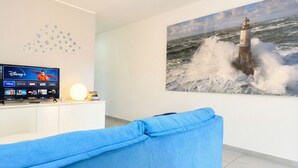 A living room with a flat-screen TV displaying Disney+ and other channels, a blue couch, and a large painting of a lighthouse in rough seas.
