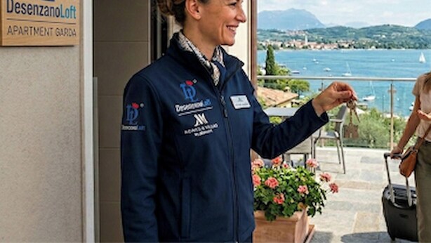 A person standing at a hotel entrance with a sign reading 'Desenzano Loft APARTMENT GARDEN'.