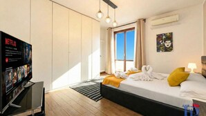 A hotel room with a bed, a TV, a window, and a painting on the wall.