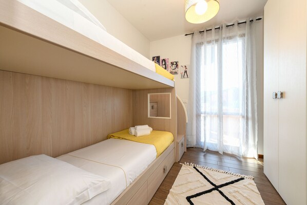 Un dormitorio con una cama litera, una cama individual, una mesita de noche, un espejo y una ventana con cortinas.
