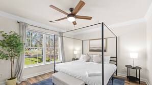 A four-poster bed with a ceiling fan, a large window with curtains, a bedside table with a lamp, and a potted plant.