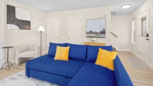 A room with a blue sectional sofa, a small white chair, a floor lamp, and a painting on the wall.