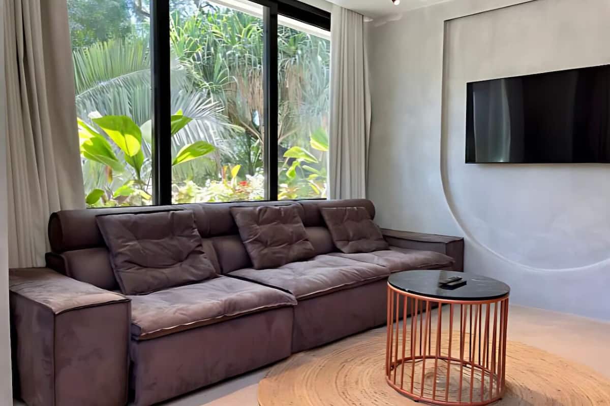 Un salon moderne avec un canapé d’angle, une table basse ronde et une grande fenêtre donnant sur un paysage verdoyant.