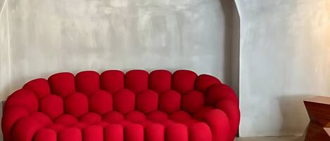 A red, circular, upholstered sofa with a unique, segmented design, placed in a room with a plain, light-colored wall and a wooden side table.