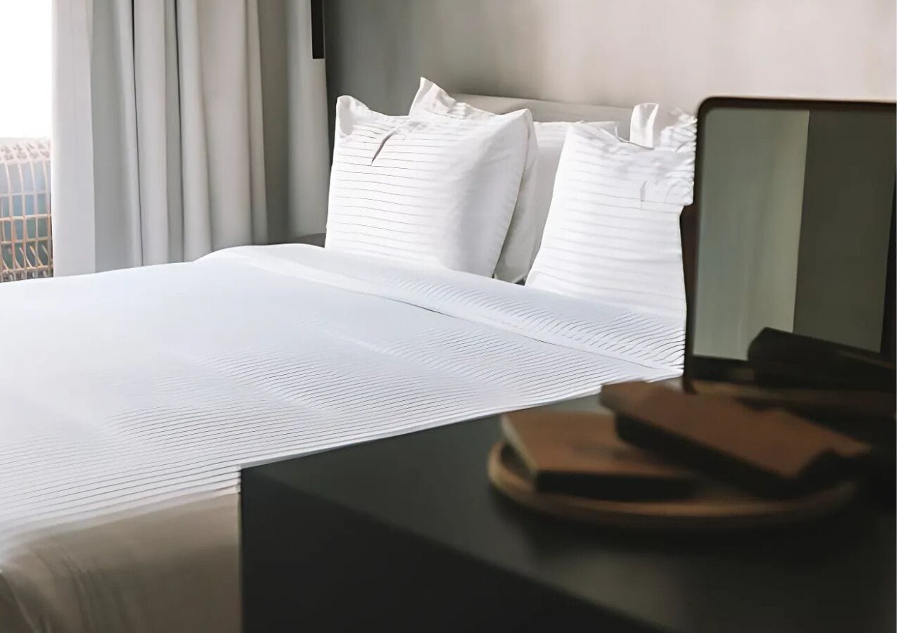 Una cama bien hecha con sábanas blancas, una mesita de noche de madera oscura con un libro y un control remoto, y una ventana con cortinas.