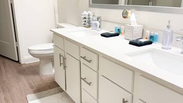 Une salle de bain moderne avec un lavabo blanc, un miroir et des armoires. On aperçoit des toilettes et une douche à l’arrière-plan.