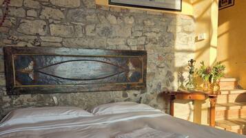 A bed with neatly folded towels, a rustic stone wall, a framed artwork, and a small wooden table with a vase.