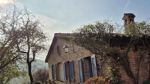 A rustic stone building with a sloped roof, a chimney, and a small tower. There is a garden with various plants and a pathway leading to the entrance. - agriturismo il melo rosso   (Fortunago)