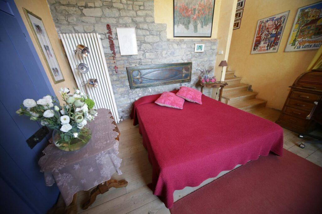 A bedroom with a stone wall, a bed with a red bedspread, a wooden dresser, and a staircase.