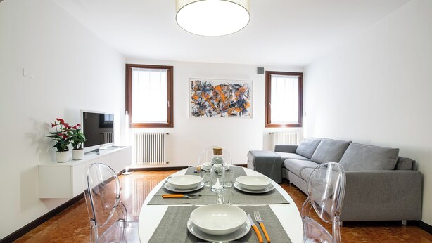 Een moderne woonkamer met een eettafel gedekt voor vier personen, een grijze bank en een groot abstract schilderij aan de muur.