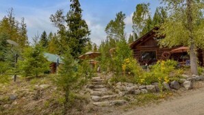 A rustic cabin with a stone pathway leading to the entrance, surrounded by dense greenery and trees. - Authentic lofted log cabin with hot tub (Kelowna)