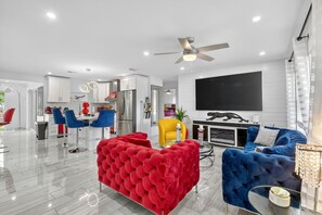 Una sala moderna con un sofá rojo y azul, un sillón amarillo y una zona de comedor con sillas azules.