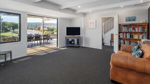 A modern living room with a large window, a flat-screen TV, a bookshelf, and a staircase leading to another level. - Spacious Family Getaway - Whangamata Holiday Home (Whangamata)