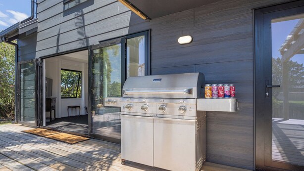 A modern outdoor kitchen with a stainless steel grill, a sliding glass door entrance, and a deck with a wooden floor. - Spacious Family Getaway - Whangamata Holiday Home (Whangamata)
