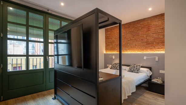 Une chambre moderne avec une grande armoire, un lit agrémenté d’oreillers à motifs, un mur en briques et une vue sur l’extérieur, visible à travers une porte verte.