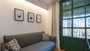 Un salon moderne avec un canapé gris, deux coussins à motifs et trois tableaux encadrés accrochés au mur. Une grande fenêtre, ornée de rideaux, offre une vue sur les bâtiments à l’extérieur.
