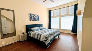 Une chambre à coucher avec un lit, un ventilateur de plafond, une fenêtre avec des rideaux et un tableau au mur.