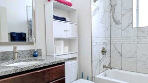 Une salle de bain dotée d’un comptoir en granit, d’un lavabo blanc, de toilettes blanches, d’un meuble-lavabo blanc avec un miroir et d’une douche aux carreaux de marbre.
