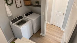 A laundry room with two washing machines, a drying rack, a wicker basket, a framed instruction sign, and a small potted plant.