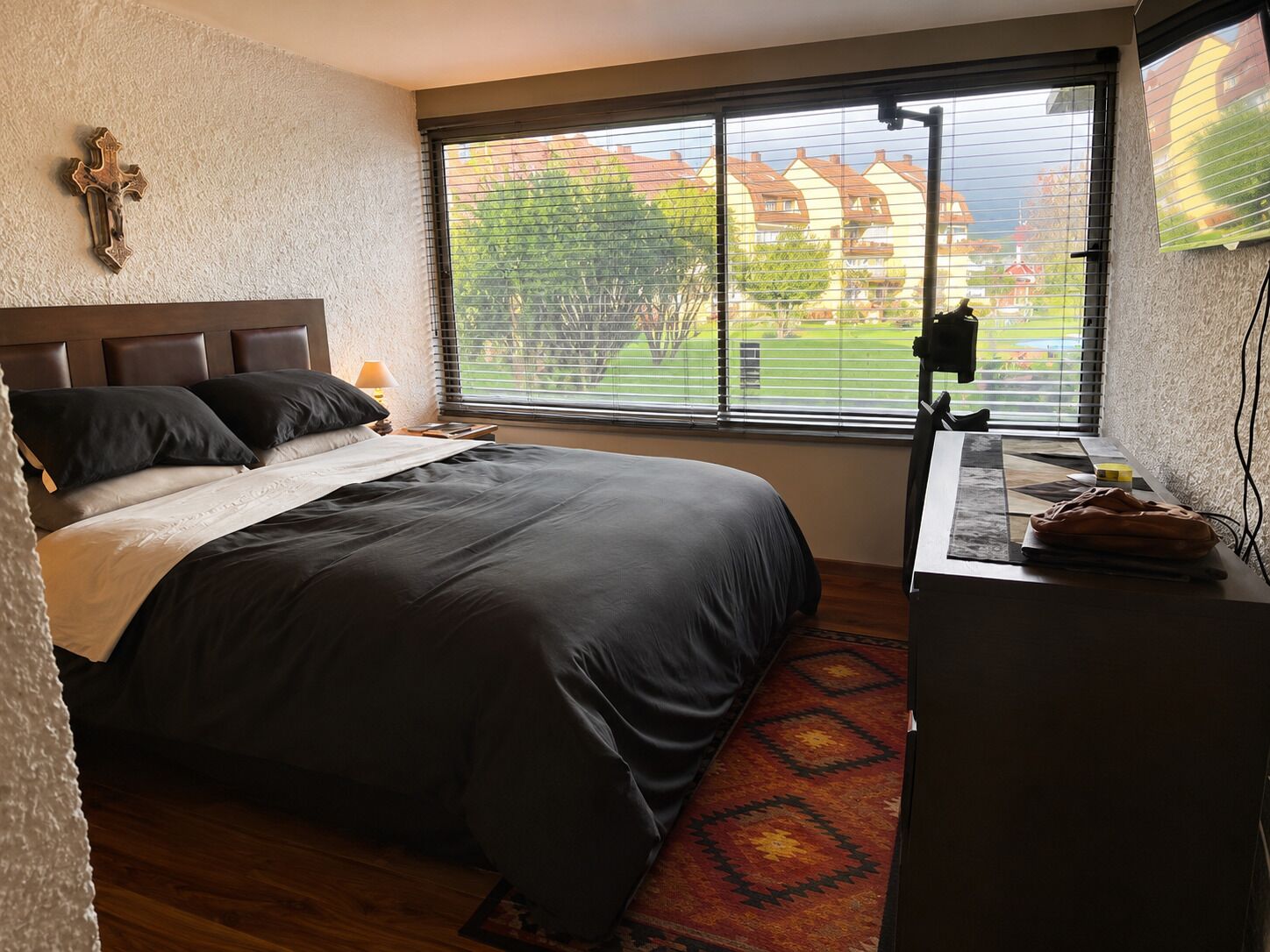 Een slaapkamer met een groot bed, een bureau met stoel, een raam met zonwering en een kruis aan de muur.