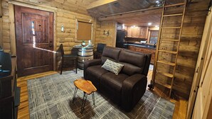 Ein gemütliches Blockhaus-Interieur mit einem Ledersofa, einem Esstisch mit Stühlen, einer Küchenzeile und einer Holzleiter.