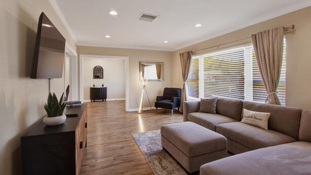 A modern living room with a sectional sofa, a flat-screen TV, a potted plant, and a wooden floor. - 4BR/3BR - Spacious Home 4 Minutes From Beach (Largo)