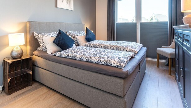 A bedroom with a bed, bedside table, lamp, artwork, window, and curtains. - Ferienwohnung in Busum (Büsum)